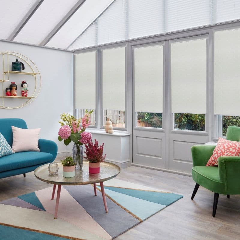 Light-colored roller blinds, lowered across tall glazed windows and doors, filtering daylight and leaving narrow lower panes exposed; bright conservatory-style living room with teal and green armchairs, coffee table, flowers.