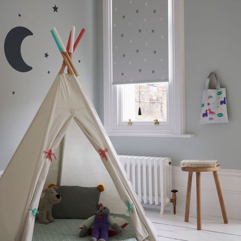 Grey roller blind with small star pattern lowered over a sash window, partially filtering daylight; canvas teepee with toys occupies foreground in a pale-grey children's bedroom, with radiator and stool.
We're Going on a Bear Hunt