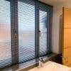 Dark horizontal Venetian blinds cover a three-panel frosted window to sill; slats slightly tilted, filtering daylight and obscuring view. Bathroom sink with chrome faucet in foreground beside wooden door.