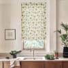 Floral-patterned roller blind covers a kitchen window, filtering light above a sink; countertop holds bowls of greens, a towel, framed sketch on wall and potted plants.