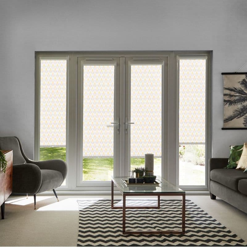Patterned roller blinds on French doors and sidelights, lowered with clear bottom strips, filtering daylight; in a bright living room with grey sofas, coffee table, chevron rug and garden view.