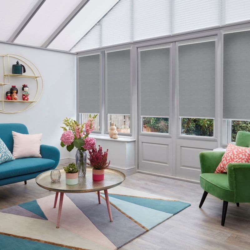Gray roller blinds cover tall conservatory windows and triangular panes, partially lowered to reveal lower glass; filtering daylight into a living room with pastel sofas, round coffee table and flowers.