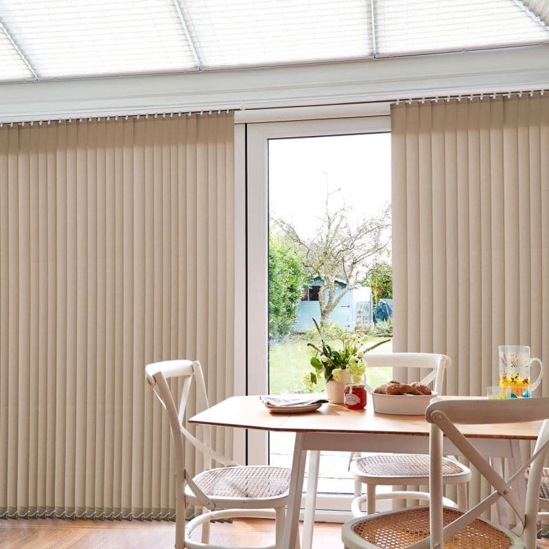 Beige vertical blinds covering a sliding glass door, mostly drawn with a central opening; filtering daylight into a dining area with wooden table, white chairs, flowers, pastries, garden beyond.