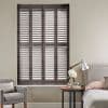 Floor-to-ceiling wooden plantation shutters covering the window, closed with angled louvers filtering daylight; set in a neutral bedroom with upholstered bed, nightstand, lamp, vase of flowers and armchair.