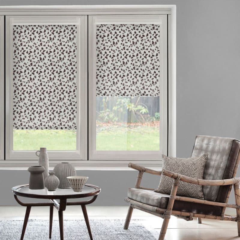 Patterned roller blinds (white with dark leaf-like shapes) lowered over the upper two-thirds of windows, filtering daylight in a grey-walled living room with rattan armchair and coffee table.