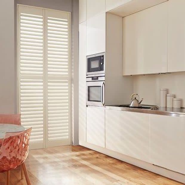 White floor-to-ceiling plantation shutters, closed and slatted, filtering sunlight and casting striped shadows across wooden floor and white cabinets; modern minimalist kitchen with built-in ovens, kettle, and pink translucent chair.