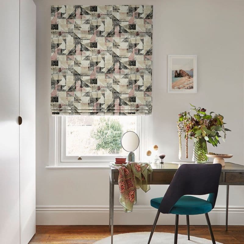Geometric-patterned Roman blind, lowered just above the sill, softly filtering daylight; a desk beneath holds a round mirror, vase of flowers and a scarf-draped surface, paired with a teal chair.
