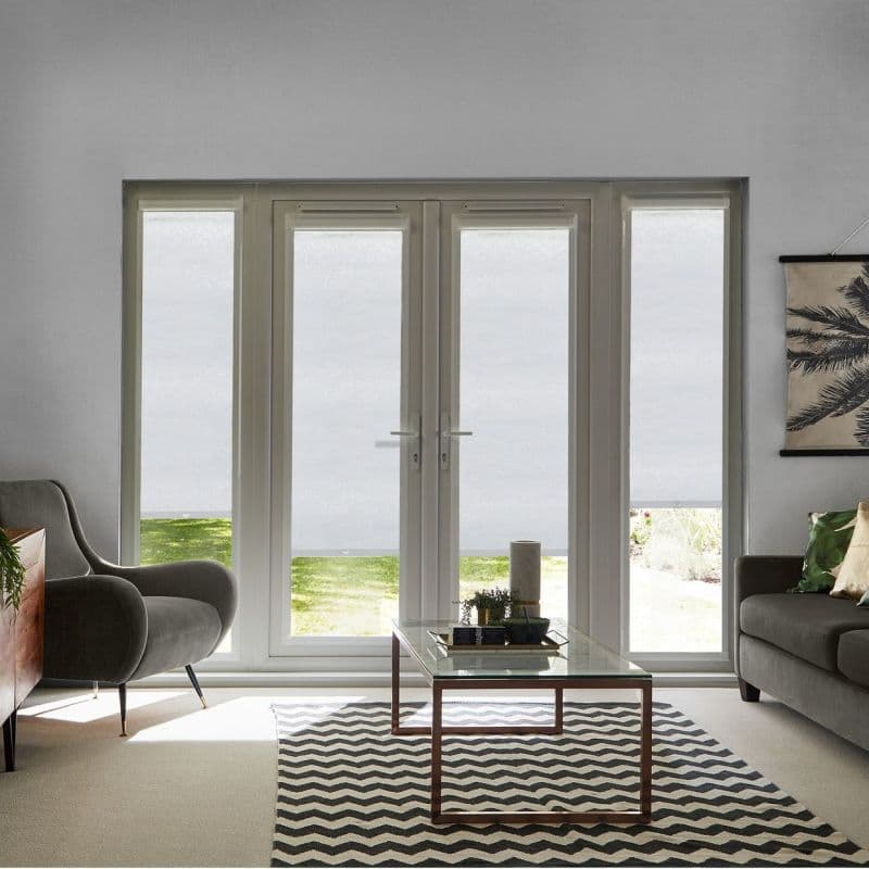 White roller blinds cover four-panel French doors and sidelights, lowered mostly with bottom gaps; filtering daylight; in a neutral living room with armchairs, glass coffee table, chevron rug, garden view.