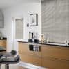 Silver horizontal (Venetian) blinds covering a large window and smaller door, slatted and partially closed to filter daylight; modern bright kitchen with light wood cabinets, black countertop, bar stools.