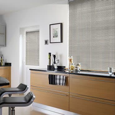 Silver horizontal (Venetian) blinds covering a large window and smaller door, slatted and partially closed to filter daylight; modern bright kitchen with light wood cabinets, black countertop, bar stools.