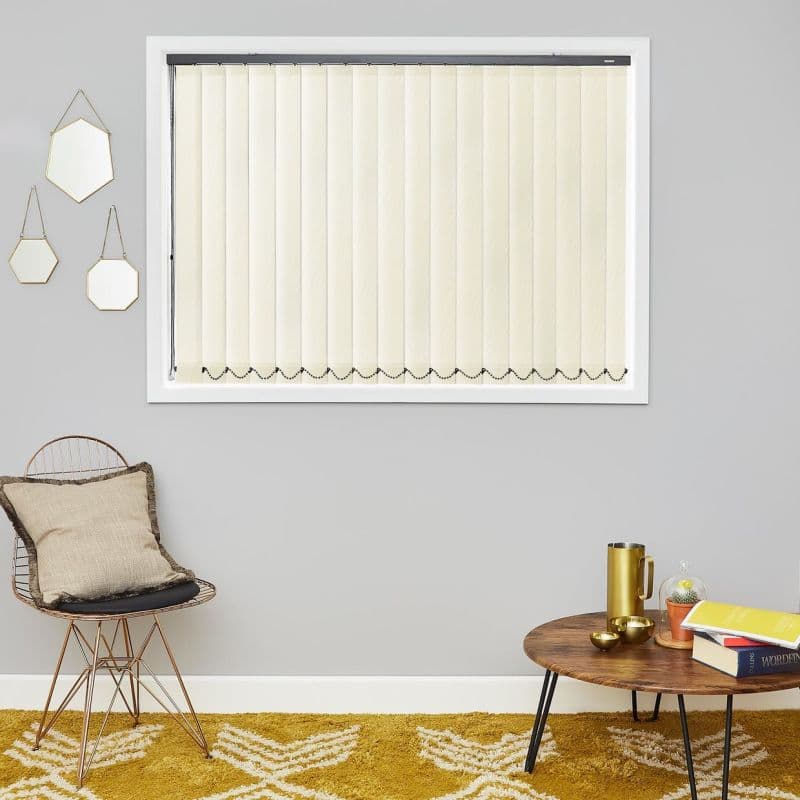 Cream vertical blinds, fully closed across a white-framed window with bead chains at the bottom, filtering daylight; living room with grey wall, wire chair, wooden coffee table and mustard rug.