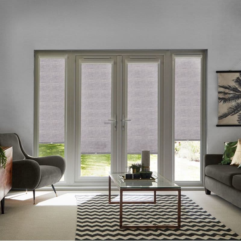 Light-gray fabric roller blinds, lowered across four-panel French doors and sidelights with small bottom gaps, filtering daylight and softening view; living room setting with armchairs, glass coffee table, chevron rug.