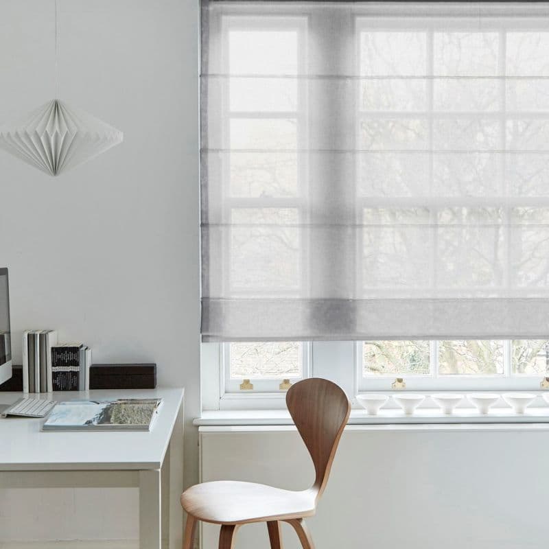 Sheer grey Roman blind lowered over a multi‑pane window, softly diffusing daylight; white desk with books and wooden chair beneath, pendant lamp left, pale minimalist room with treed view.