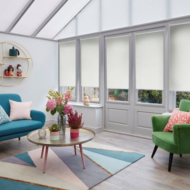 White roller blinds across tall conservatory windows and door, lowered midway and filtering light; bright modern sitting room with teal sofa, green armchair, coffee table, flowers and pastel geometric rug.