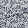 Light gray woven fabric with navy embroidered fern-like leaf pattern, folded in the center creating overlapping layers; close-up, soft diffuse lighting reveals textile texture and stitch detail.