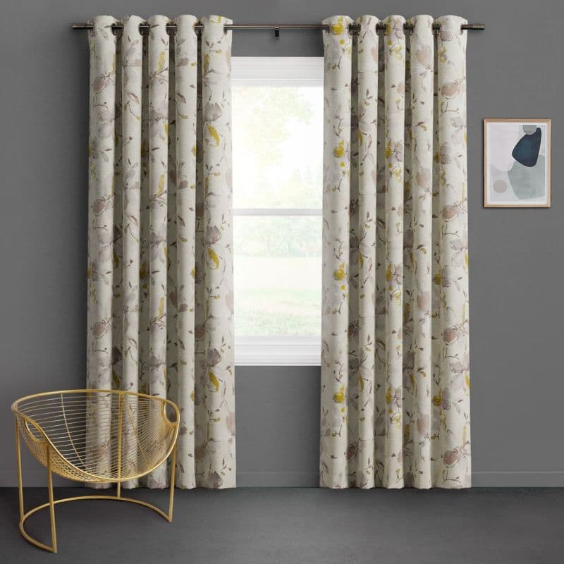 Grommet-top floral curtains, cream with muted beige and yellow blossoms, hang in pleats framing a central window, filtering soft daylight in a gray-walled room with a gold wire chair.