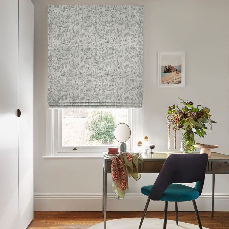 Floral-patterned Roman blind lowered over a sash window, gently filtering daylight; above a wooden desk with mirror, teal chair and vase of flowers in a bright home office.