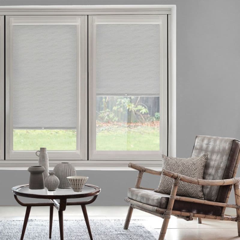 Roller fabric blinds, light grey and textured, partially lowered over twin windows, filtering daylight; modern living room with wooden coffee table and ceramics, rattan-framed leather chair, and a garden view.