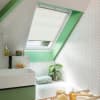 White pleated skylight blind, partially lowered over a roof window, filtering soft daylight into a bright attic bathroom framed in mint-green trim with white herringbone tiles, sink, tub, plant.
