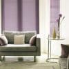 Purple horizontal blinds covering tall windows, fully closed and filtering soft light; a gray upholstered sofa with pillows faces a glass side table holding vases, in a lit living room.