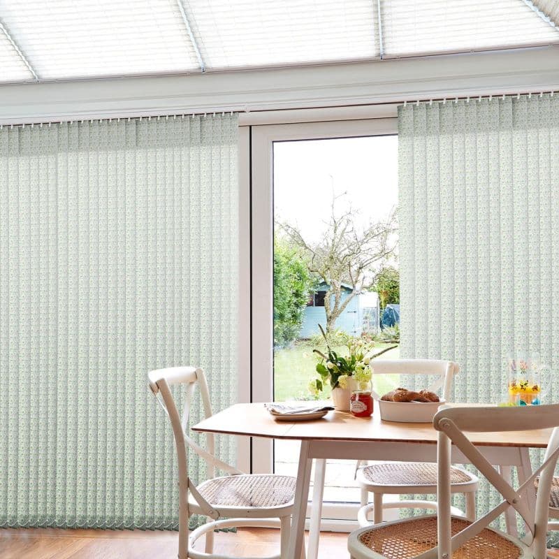 Pale green patterned vertical blinds covering a sliding glass door, partially parted at center and filtering soft daylight; small dining table with chairs, vase and pastries, backyard garden beyond.