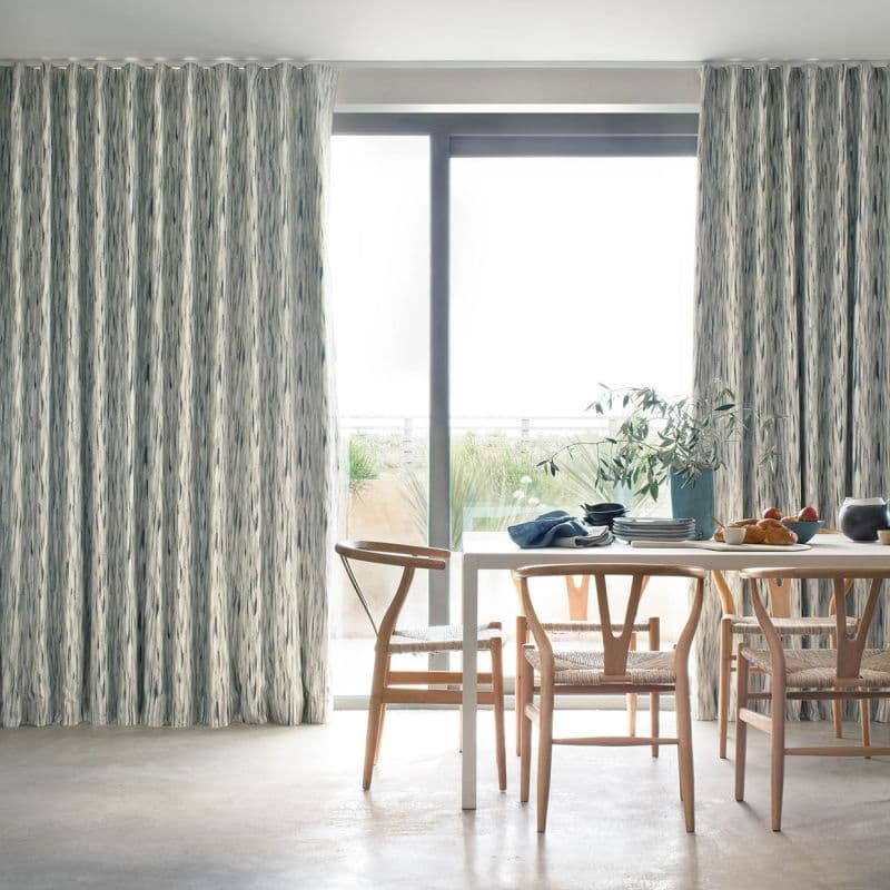 Full-length, pleated patterned curtains (grey‑white vertical brushstrokes) partially drawn, framing a sliding glass door and filtering bright daylight; dining table with wooden chairs, tableware, plant, balcony view.