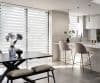 Striped dual‑layer roller blinds across full-height windows, alternating sheer and opaque bands, filtering sunlight and casting horizontal shadows across a modern open-plan kitchen-dining area with marble island and bar stools.