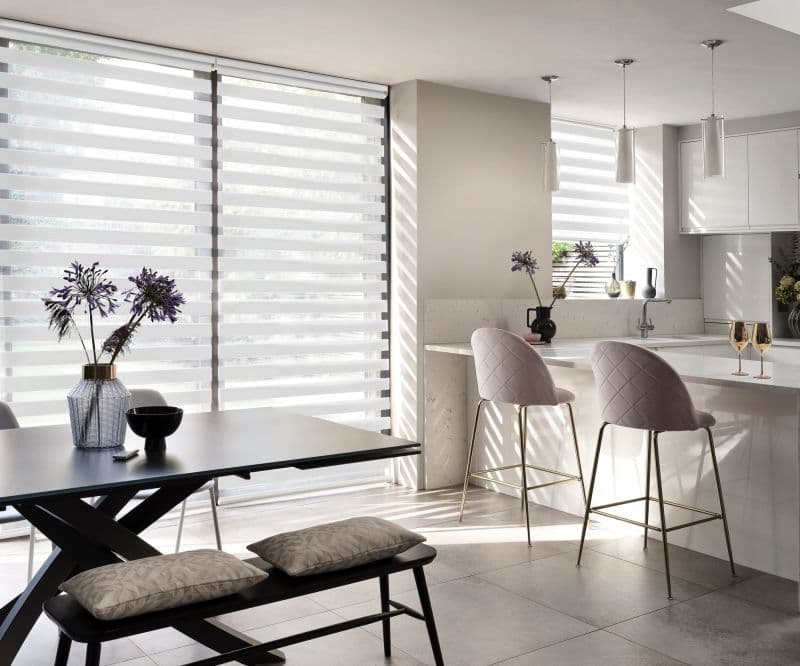 Striped dual‑layer roller blinds across full-height windows, alternating sheer and opaque bands, filtering sunlight and casting horizontal shadows across a modern open-plan kitchen-dining area with marble island and bar stools.