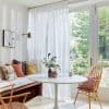 Sheer white floor-to-ceiling curtains, pleated and drawn across glass doors, filtering soft daylight; bright dining nook with round white table, wooden chairs, cushioned bench, and leafy garden view.
