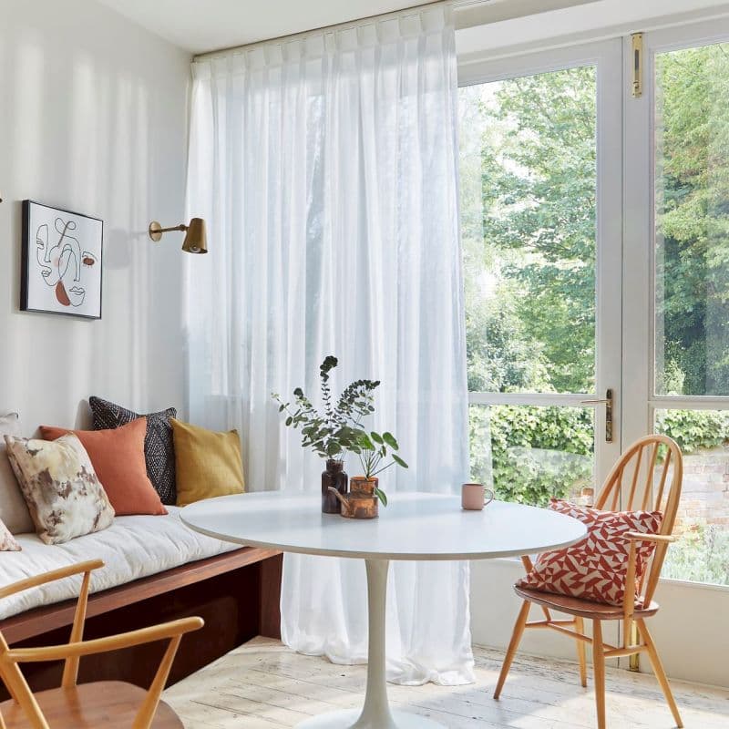 Sheer white floor-to-ceiling curtains, pleated and drawn across glass doors, filtering soft daylight; bright dining nook with round white table, wooden chairs, cushioned bench, and leafy garden view.