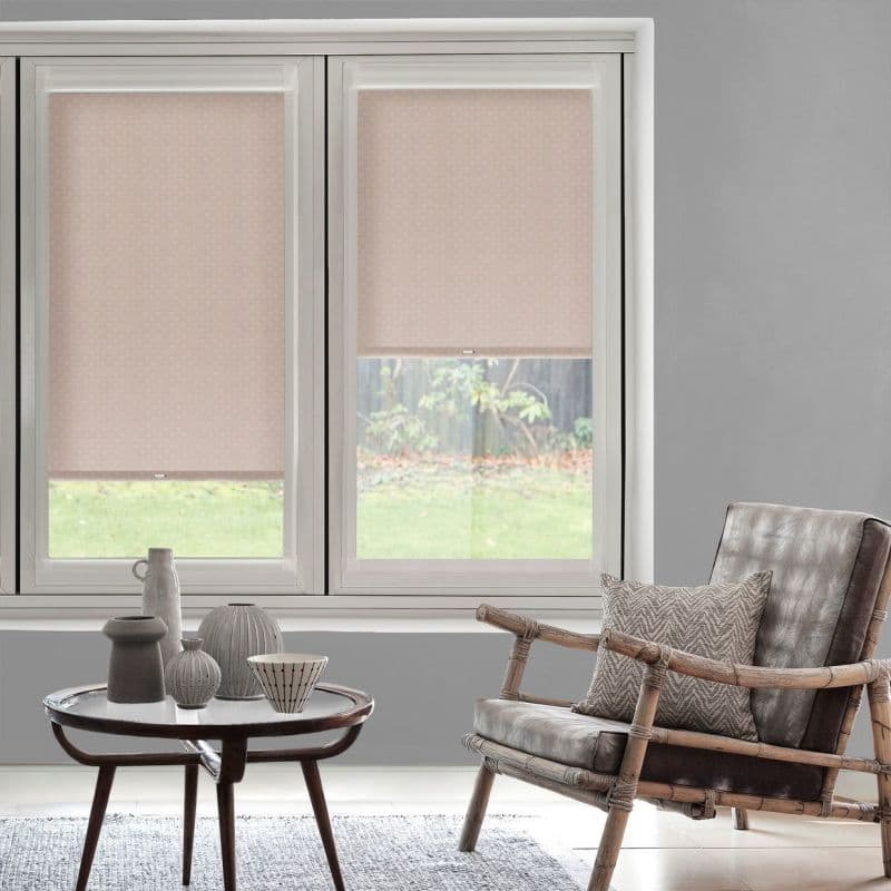 Pink patterned roller blinds: left fully lowered, right lowered mid-window; they filter light, partially blocking the view. Context: gray living room with rattan armchair, side table, soft daylight, garden beyond.