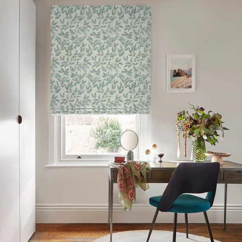 Roman blind with green leafy pattern, lowered partway, filtering daylight; a desk and teal chair hold a mirror, scarf, and vase of flowers in a sunlit home workspace.