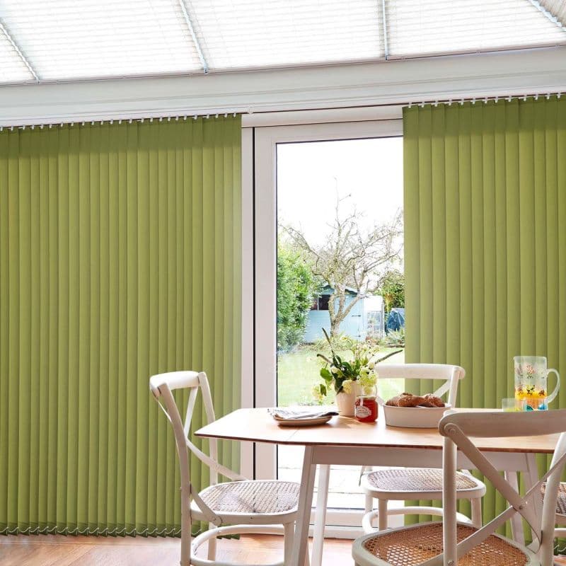 Green vertical blinds covering a sliding glass door, partially parted at center; filtering daylight into a bright dining nook with white chairs, wooden table, and a small garden visible outside.