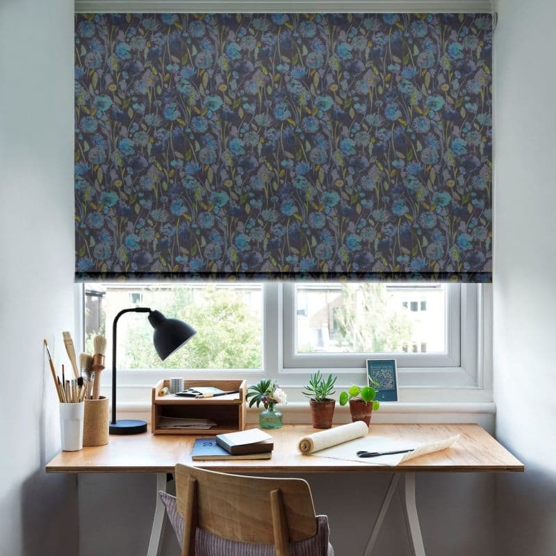 A dark floral roller blind, lowered across the window with blue flowers, filtering daylight; above a wooden desk with lamp, paintbrushes, potted plants and notebooks, light visible through a gap.