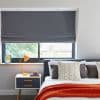 Grey fabric Roman blind lowered and mostly closed, filtering light with a small gap; bedroom interior with bed, patterned cushions, orange throw and bedside table with lamp.
12:01