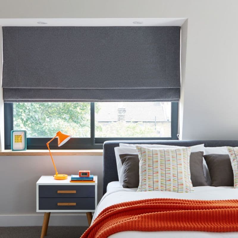 Grey fabric Roman blind lowered and mostly closed, filtering light with a small gap; bedroom interior with bed, patterned cushions, orange throw and bedside table with lamp.
12:01