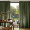 Green pleated floor-length curtains parted to reveal a glass sliding door, filtering soft daylight. Dining table set with pancakes and fruit, pendant lamp overhead, backyard garden and trees visible.