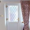 Floral pleated curtains, drawn right and tied back with matching rope, partially covering a white diamond-leaded glass door with brass handle; soft daylight filters into the entry hall.