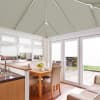 Pale pleated cellular blinds cover the conservatory roof, upper windows and French doors, honeycomb-textured and partially lowered, filtering bright daylight into a compact dining area overlooking a paved garden.