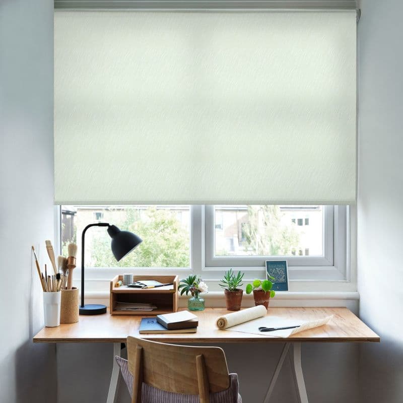A pale patterned roller blind, fully lowered across a double window, gently filtering daylight; a wooden desk workspace below with lamp, brushes, notebooks, potted succulents and a suburban view.