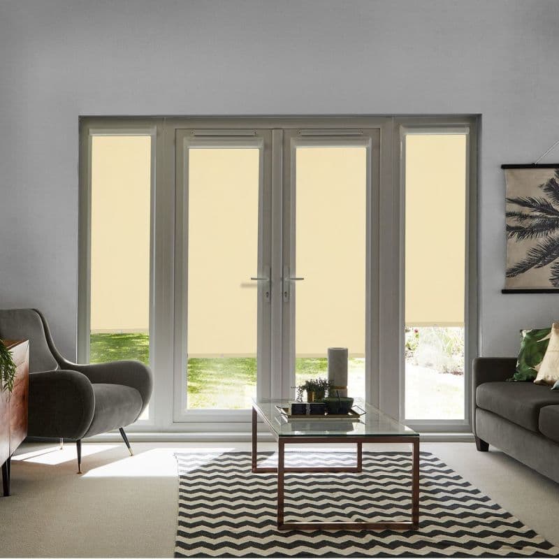 Beige roller blinds covering tall French doors and flanking sidelights, lowered to mid-height and filtering daylight; modern living room with gray armchairs, glass coffee table, chevron rug and garden view.