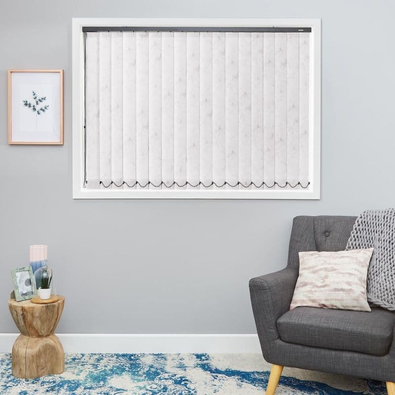 White marbled vertical blinds cover the window—closed, aligned slats with beaded scalloped weights and side control chain—filtering daylight in a grey-walled living room with armchair, side table, blue rug.