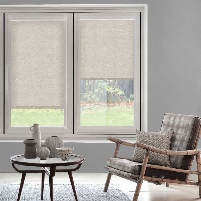 Beige fabric roller blinds cover twin windows—left fully lowered, right partially raised—filtering soft daylight. Modern gray living room with rattan armchair, patterned cushion, round coffee table and grassy backyard.