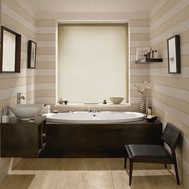 Beige roller blind over window — fully lowered, diffusing soft daylight — above a dark-wood inset bathtub in neutral striped-wall bathroom with vessel sink, floating shelf, chair and warm tile flooring.