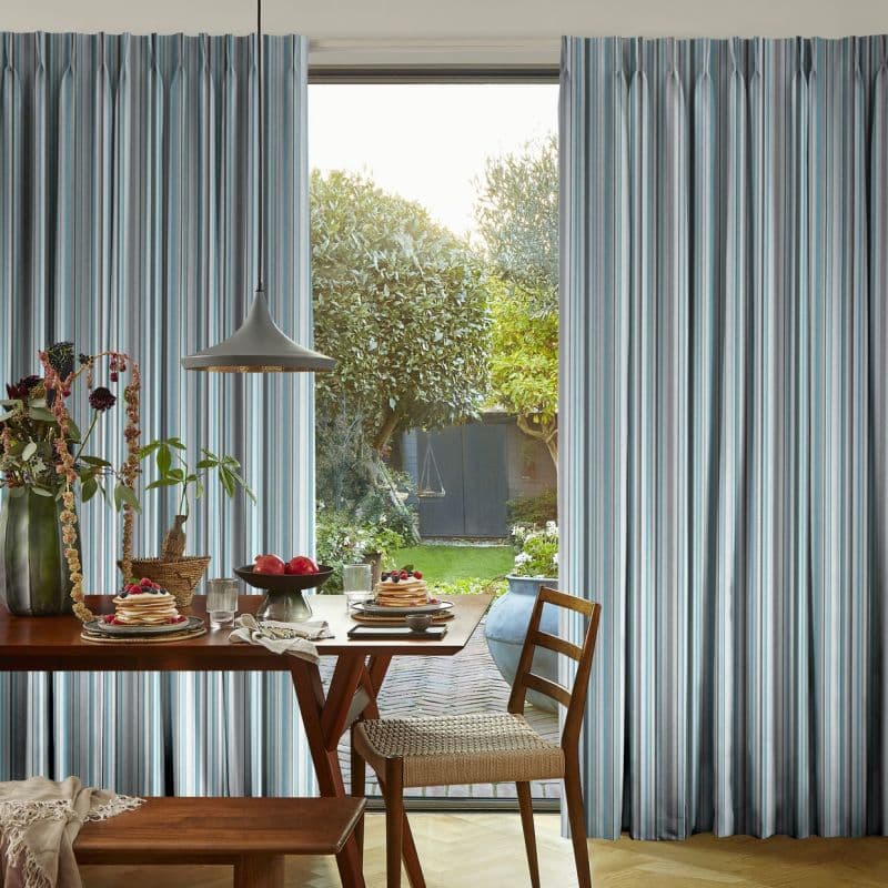 Striped, pinch-pleat floor-length curtains (blue, gray, white) parted at center, framing and softening light; a dining table with pendant lamp, chairs, and breakfast faces a sunlit backyard garden.