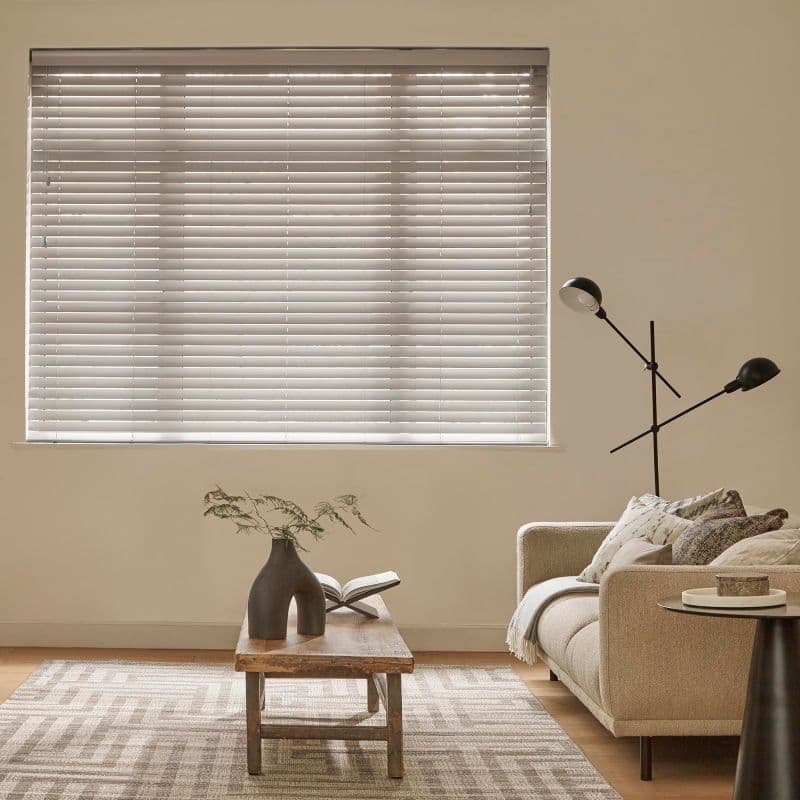 Horizontal Venetian blinds covering a large rectangular window, closed and filtering soft daylight through thin slats; living room with beige sofa, wooden coffee table, lamp, sculptural vase and open book.