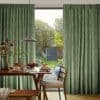 Green textured pleated curtains, parted centrally and hanging floor-to-ceiling, partly open filtering daylight; in a dining area with wooden table set for breakfast, pendant lamp, and sunlit garden view.