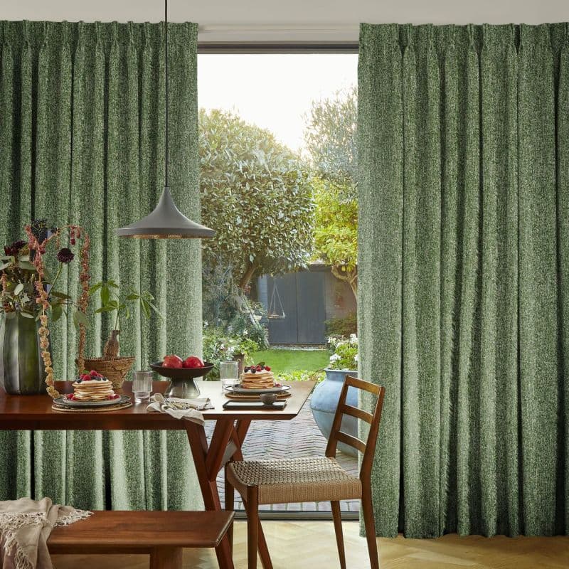 Green textured pleated curtains, parted centrally and hanging floor-to-ceiling, partly open filtering daylight; in a dining area with wooden table set for breakfast, pendant lamp, and sunlit garden view.
