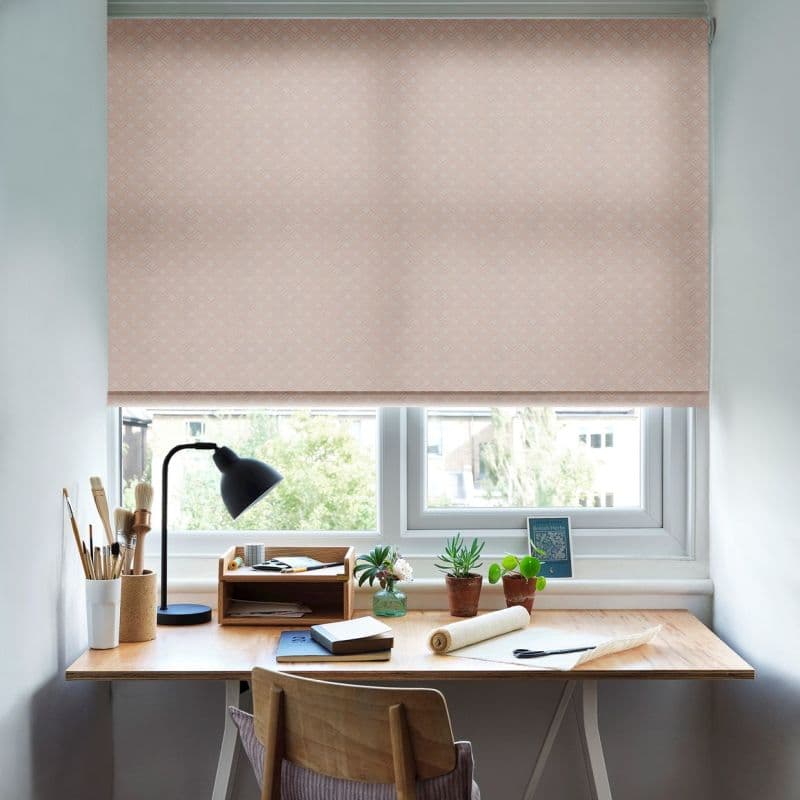 Pink patterned roller blind, fully lowered with subtle diamond texture, filtering soft daylight over a wooden home desk with lamp, art brushes, potted succulents, notebooks and suburban window view.