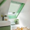 Pleated white skylight blind, partially lowered, filtering soft daylight; placed in a green-painted roof window recess above a bathtub in a bright attic bathroom with white herringbone tiles.